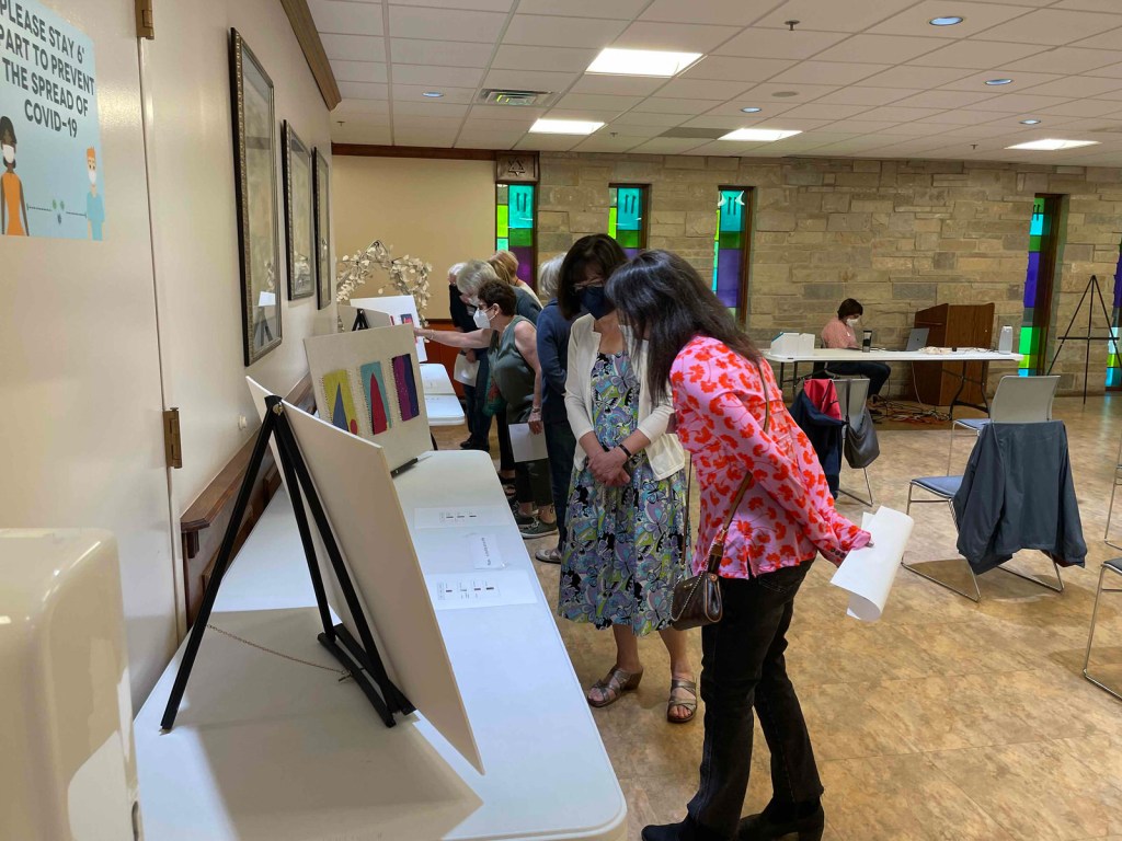 Standing people looking at tapestries displayed on easels at tables