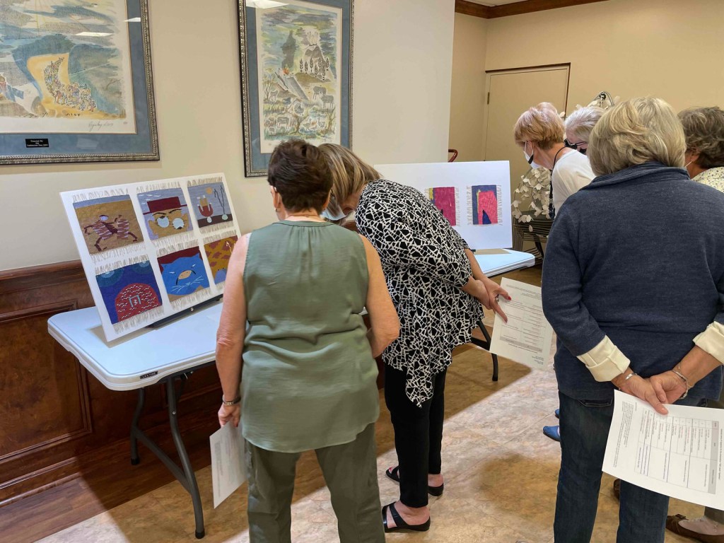 Standing people looking at tapestries displayed on easels at tables