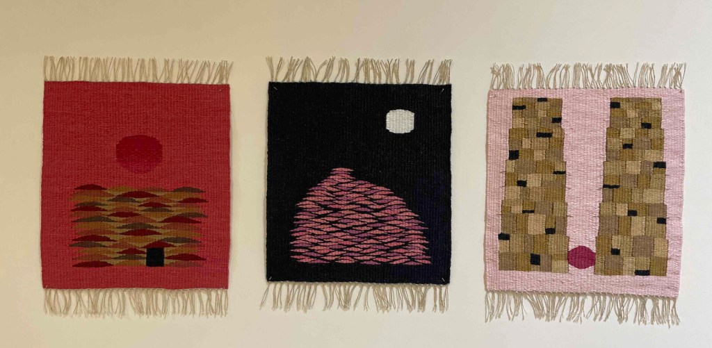 Photo of three small tapestries mounted in shades of red, tan, pink, and black