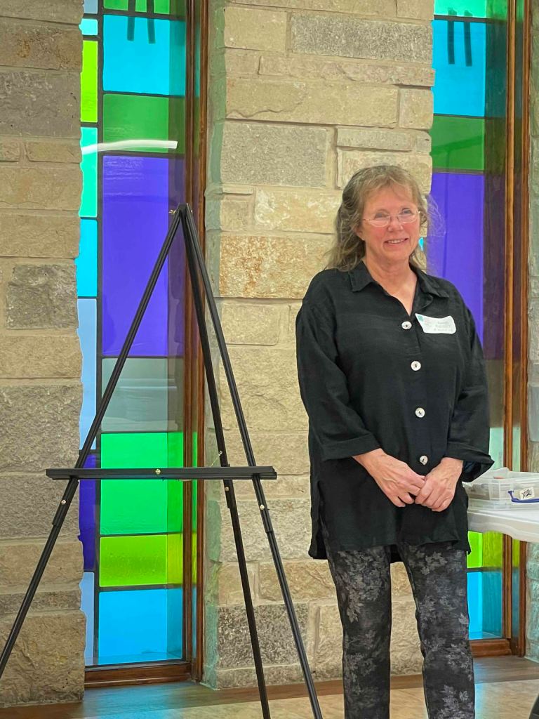 Photo of Berit Engen standing in front of a stone wall with multicolored stained glass panels and an easel to her right