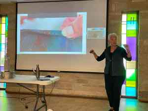Photo of women standing in front of projection screen that shows a picture of someone hemming handwoven cloth.