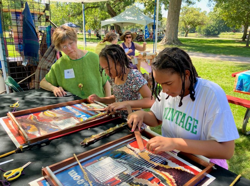 three people sitting at a table; two young people are weaving on frame looms as an adult looks on.