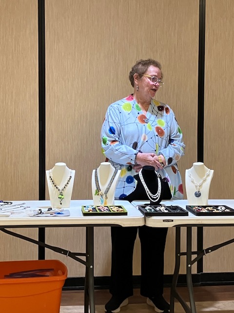 Woman standing behind jewelry display stands with necklaces made using chainmaille technique, beads, and glass focal features.