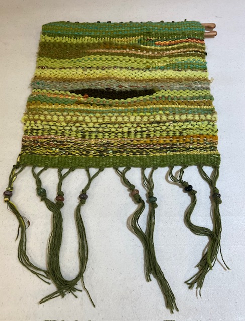 Fringed wall hanging woven in shades of green, yellow, brown, and black, with beaded fringe, mounted on a flat weaving shuttle at the top hem.