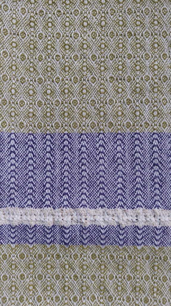 Close-up of patterned towel in shades of green, blue and cream. Pattern incorporates circles, curves, and a thin lace stripe in the blue section.