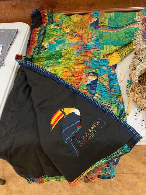 a vibrant kimono-style coat woven in shades of yellow, green, black, red, aqua, and orange, laying on a table. One corner is folded back to reveal an interior label featuring a fabric-printed toucan and the text "It's a Jungle Out There."