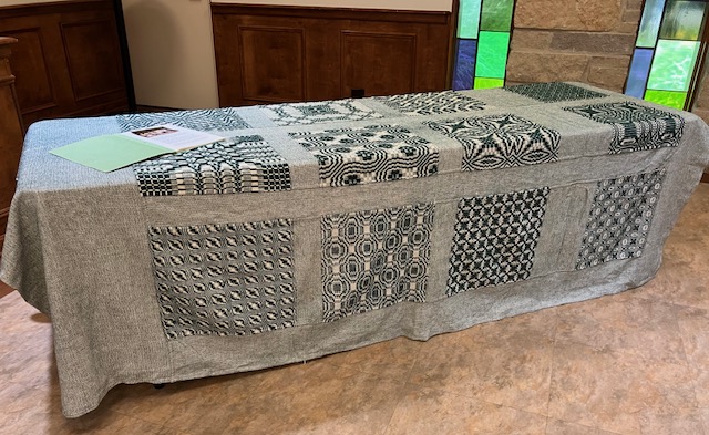 Large coverlet featuring patterned woven blocks, woven in shades of dark green and cream.