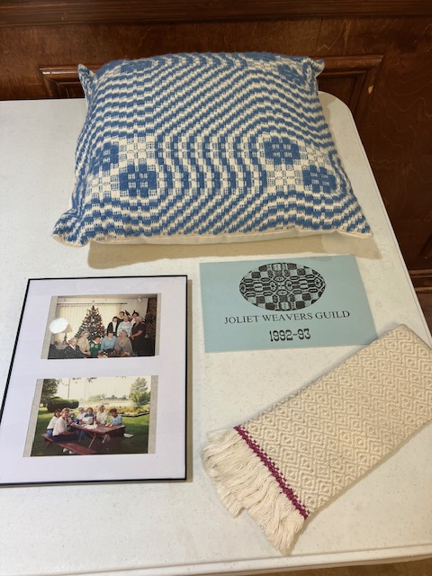 An overshot pillow in shades of blue and cream, pictures of people around a Christmas tree and a picnic table, and a handwoven fringed fingertip towel, all on a table.