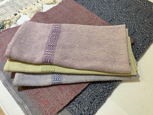 Three folded towels woven with a patterned design across one end. One towel is woven in shades of purple and cream, one is woven in shades of light green and cream, and one is woven in shades of powder blue and cream.