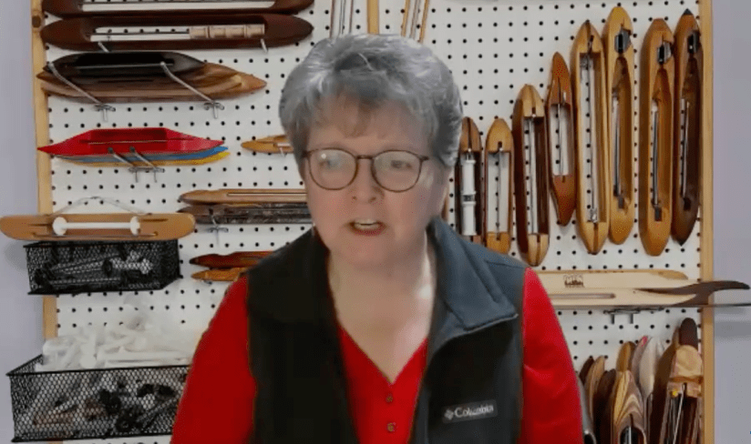 Woman wearing glasses, a red shirt, and black vest, sitting in front of a pegboard with multiple weaving shuttles attached to it