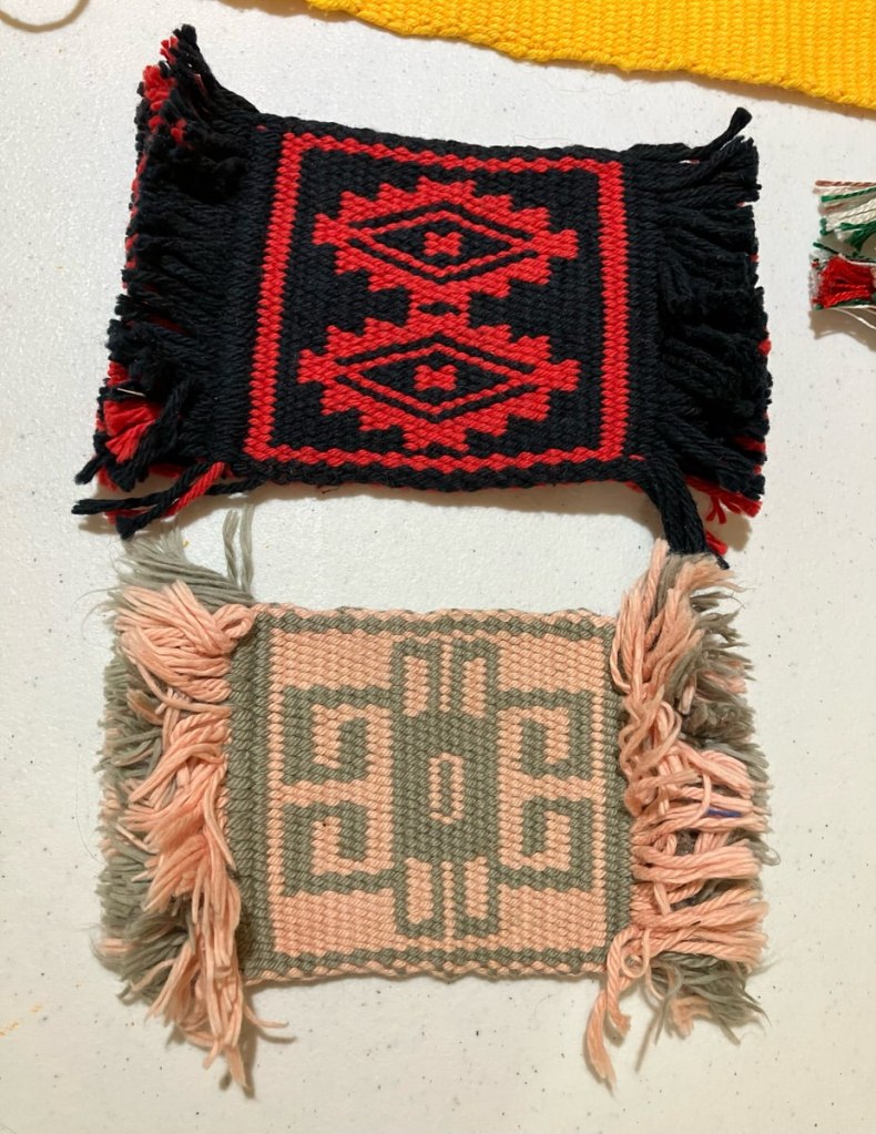 Two woven patterned mug rugs; one is woven in shades of red and black and the order in shades of peach and pale green