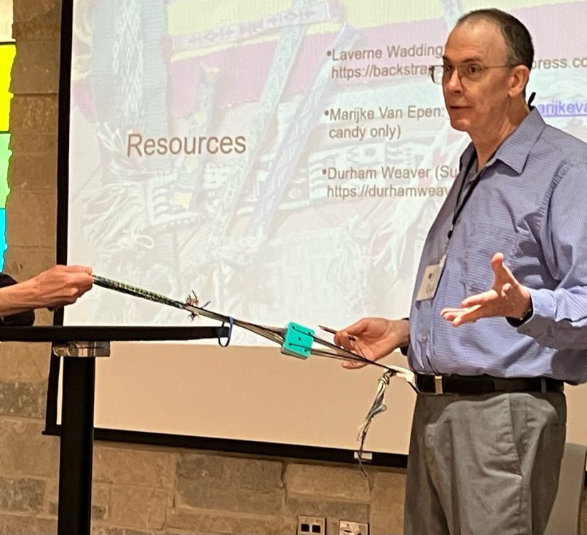 Man standing to demonstrate the use of a backstrap weaving set up using tablets (cards).