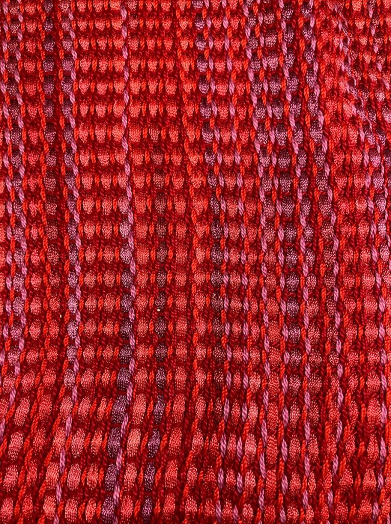 Detail of a plain woven scarf in various shades of red, purple, and pink.