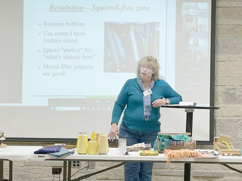 Woman standing in front of a projection screen and behind a table with weaving paraphernalia, while talking.