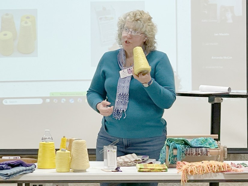 Woman standing in front of a projection screen and behind a table with weaving paraphernalia, holding a cone of yellow yarn while talking.