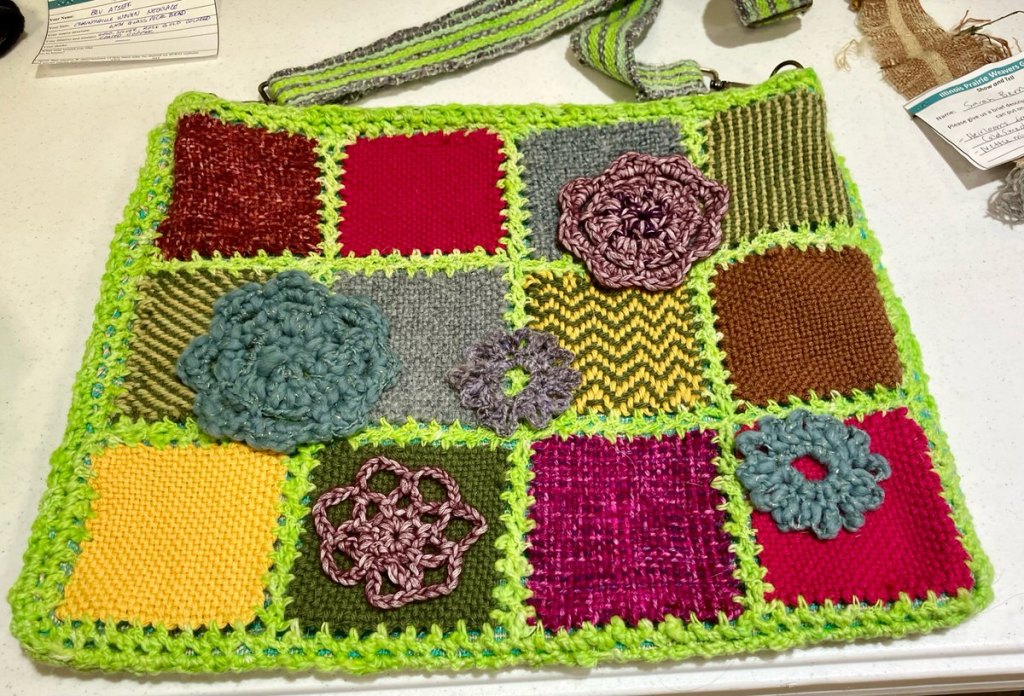 Bag made with pin woven squares in various colors, joined with a fluorescent green crochet stitch, crochet flowers appliqued. Woven matching strap.