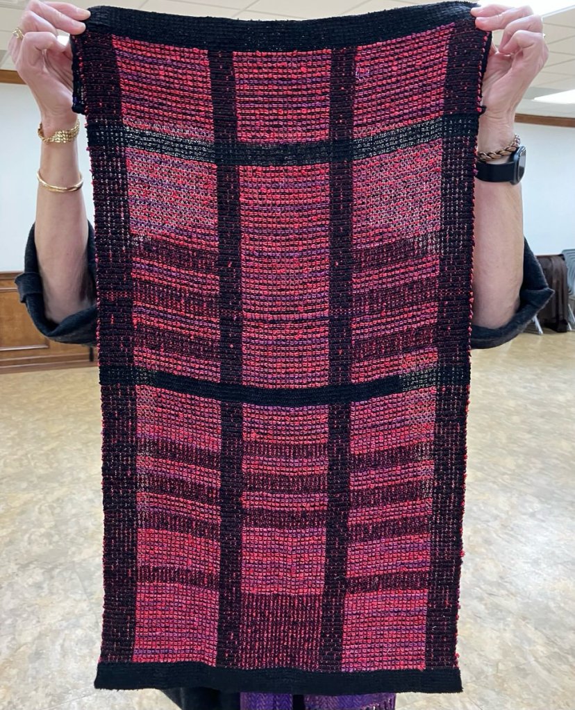 Woman behind a small wall hanging that has a black border with some black, pink, and purple yarn used for stripes.