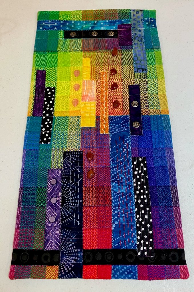Multi-colored narrow strip of twill with a few commercial fabric appliqued to it and some leaf shaped beads.
