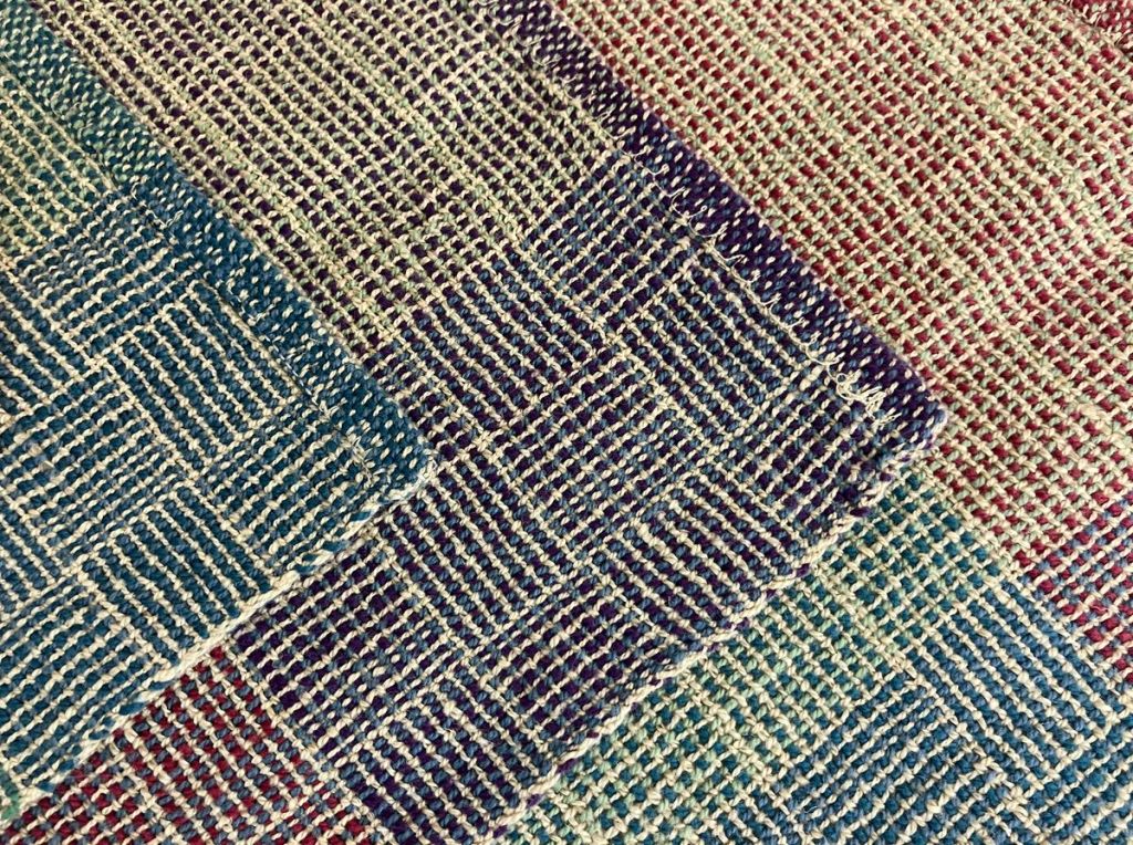 Closeup view of three patterned placemats woven in shades of teal, green, dark red, and purple