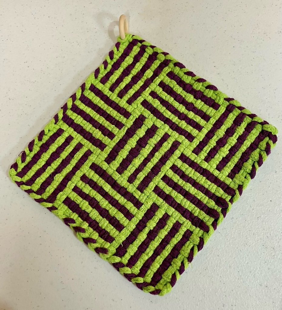 Square patterned potholder woven in shades of light green and brown with a circular wooden hanging loop in one corner