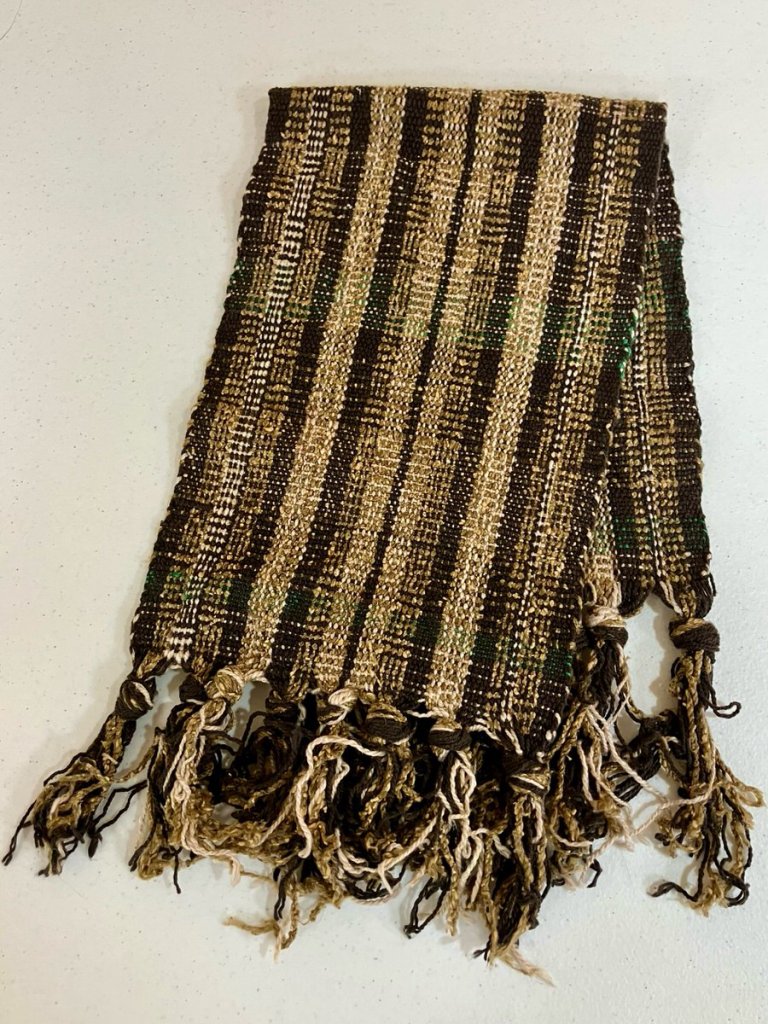 Folded, fringed cloth woven in shades of tan, black, and brown with a predominant warp-wise stripe and subtle weft-wise patterning