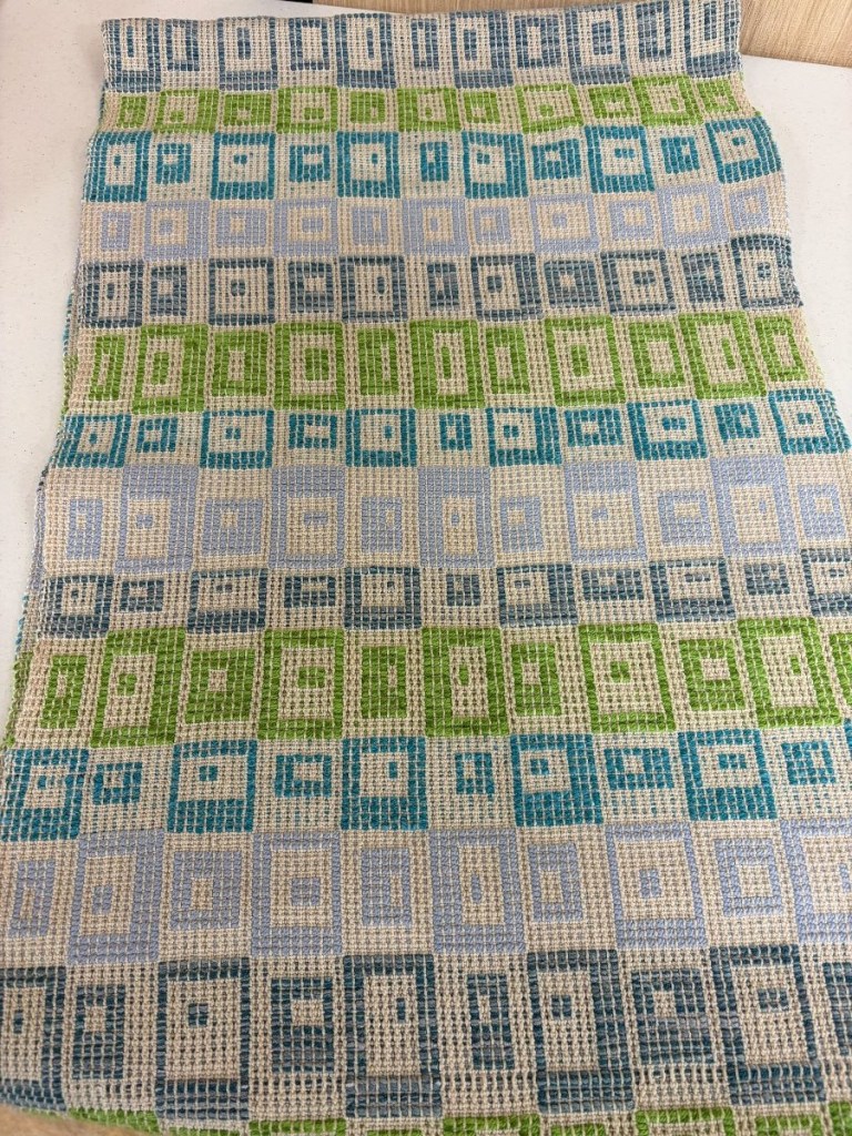 Table runner woven in a geometric pattern of rectangles in shades if blue, teal, and green on a cream-colored background