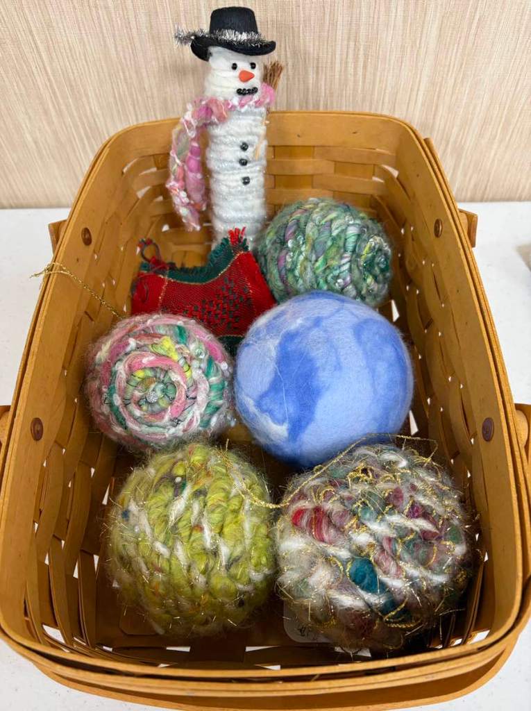 Collection of five round Christmas ornaments made with handspun yarn, and felted wool; a cylindrical snowman wearing a hat; and a small handwoven fabric swatch in shades of red and green; all in a rectangular basket