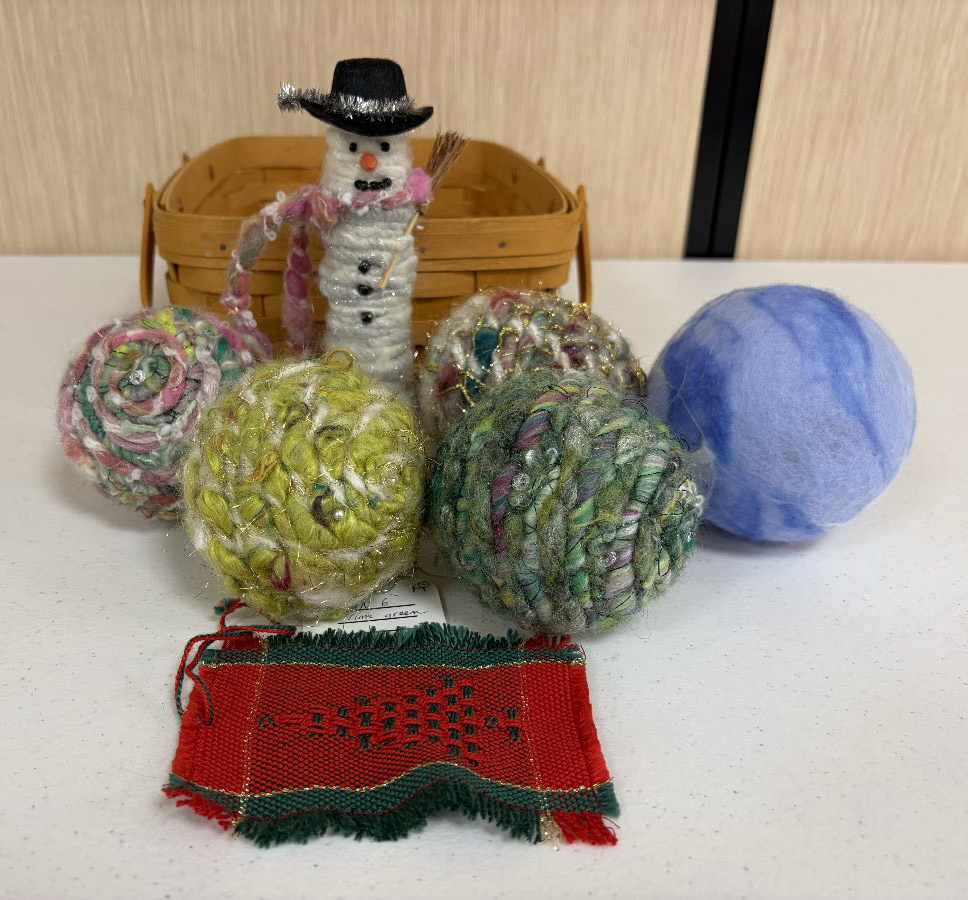 Close-up of collection of five round Christmas ornaments made with handspun yarn, and felted wool; a cylindrical snowman wearing a hat; and a small handwoven fabric swatch in shades of red and green