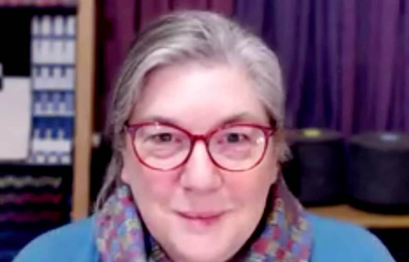 White woman with gray-dark hair and dark red eyeglasses, wearing a multi-color cowl scarf and light blue top