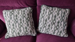 Two patterned square pillows, in shades of gray and white, positioned side-by-side on a purple velour couch/sofa