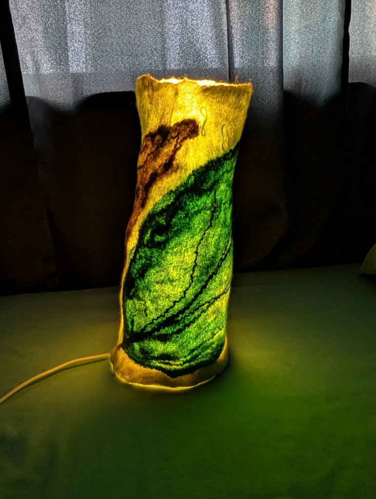 Luminary 2. Lit. Off-white tall columnar luminary with a green leaf design.