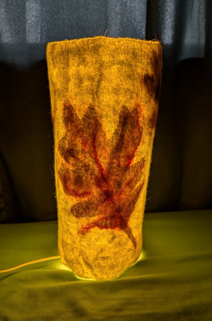 Luminary 3. Lit. Off-white tall columnar luminary with an oak leaf design.