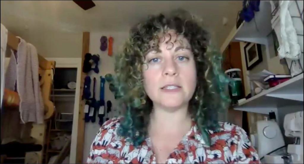 White woman with curly hair and print shirt in a fiber studio setting with a loom, sewing machines, and other fiber art tools and supplies in the background.