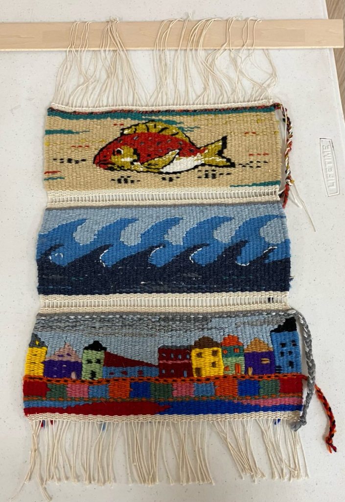 Three tapestry strips using the same warp. Top: a fish. Middle: light and dark blue waves. Bottom: city scene.