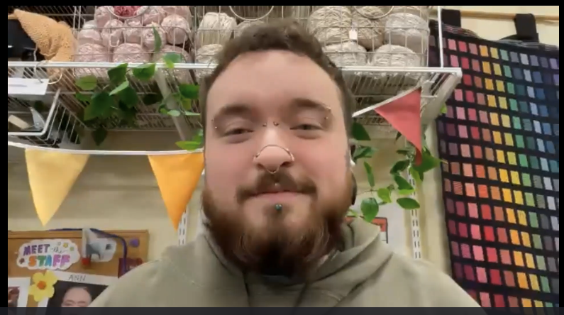 Dark-haired white man with a beard and various facial piercings, sitting in front of a wall with warn balls and a multi-colored fiber wall hanging.