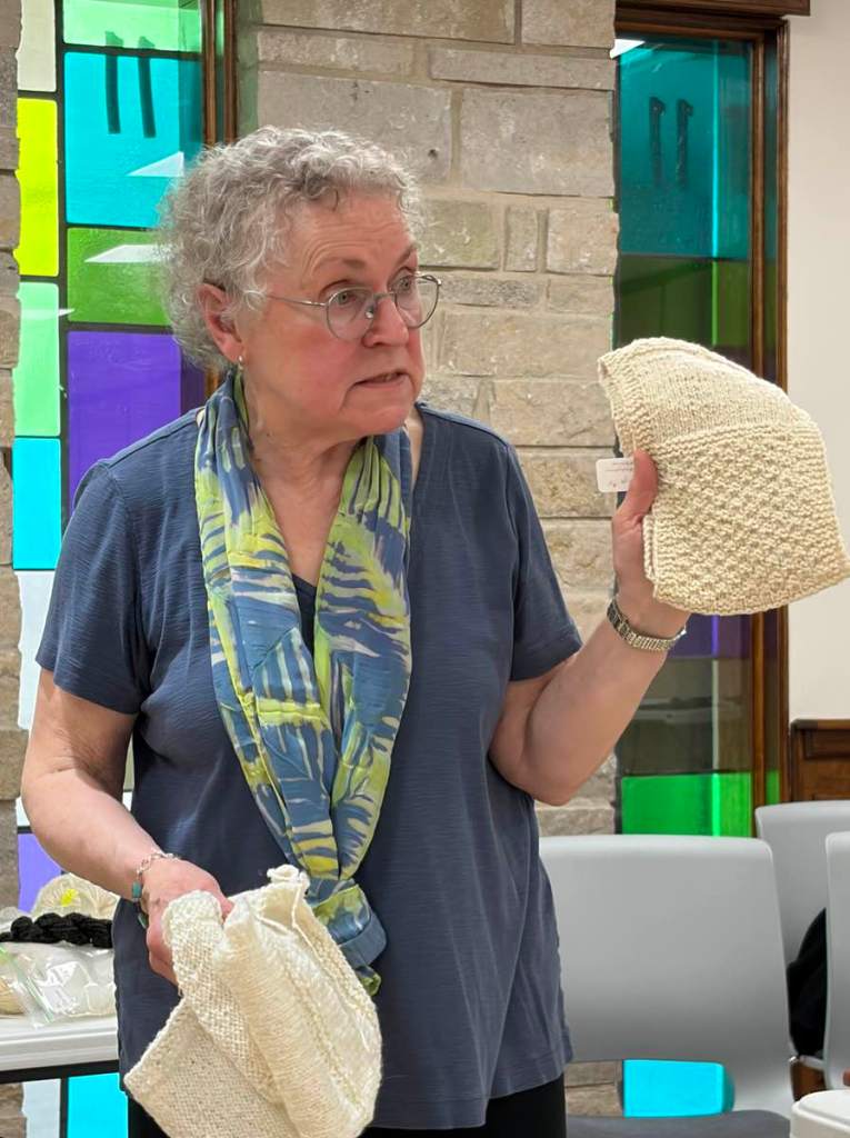 Older white woman wearing glasses, blue shirt, and blue and green scarf holds two knitted items in cream-colored wool.
