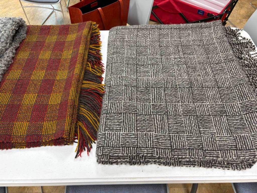 Two fringed blankets folded on a table. The blanket on the left has a checked-type pattern in shades of brick red, gold, and brown. The blanket on the rights has a "log cabin" color-and-weave pattern in shades of black and white.