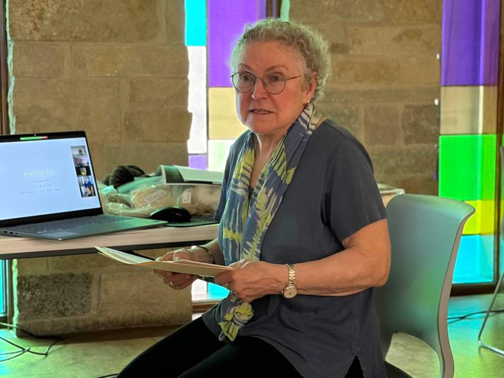 Older white woman wearing glasses, blue shirt, and blue and green scarf, holding a file folder, seated in front of a laptop computer, answers an audience question.