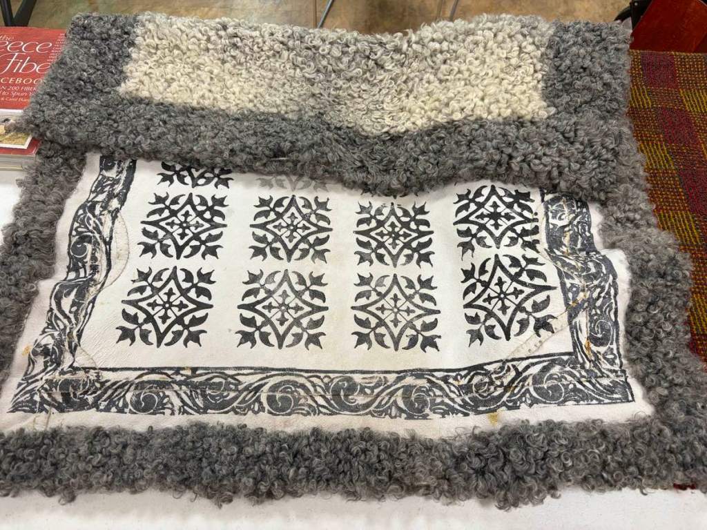 A blanket-like coverlet with two shades of wool shearling (gray and tan) on one side and a stamped gray and white design on the other.