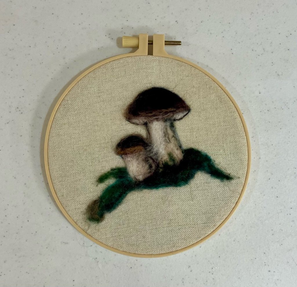 A large and small mushroom felted onto a piece of linen, in an embroidery hoop frame.