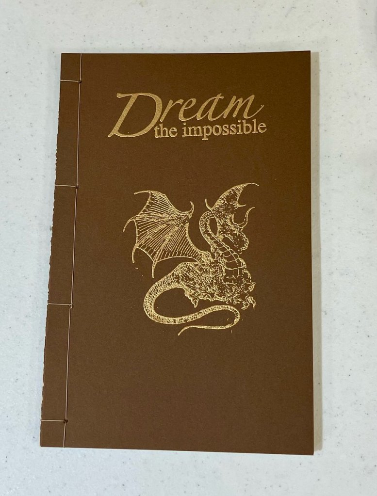 David L. - Brown handmade book with the title "Dream the impossible" at the top and a dragon underneath done in goldwork.