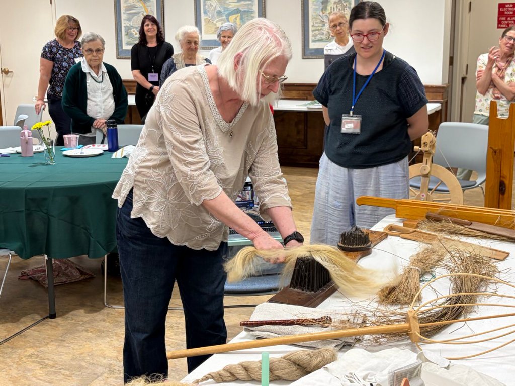 Woman standing at a table, drawing flax through a spikey implement to reveal spinnable flax fiber.