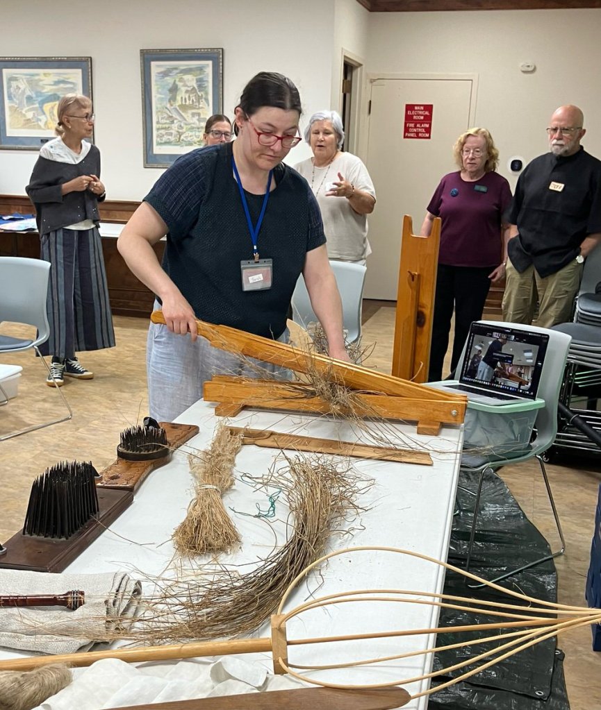 Woman standing at a table using a wooden implement to process flax.