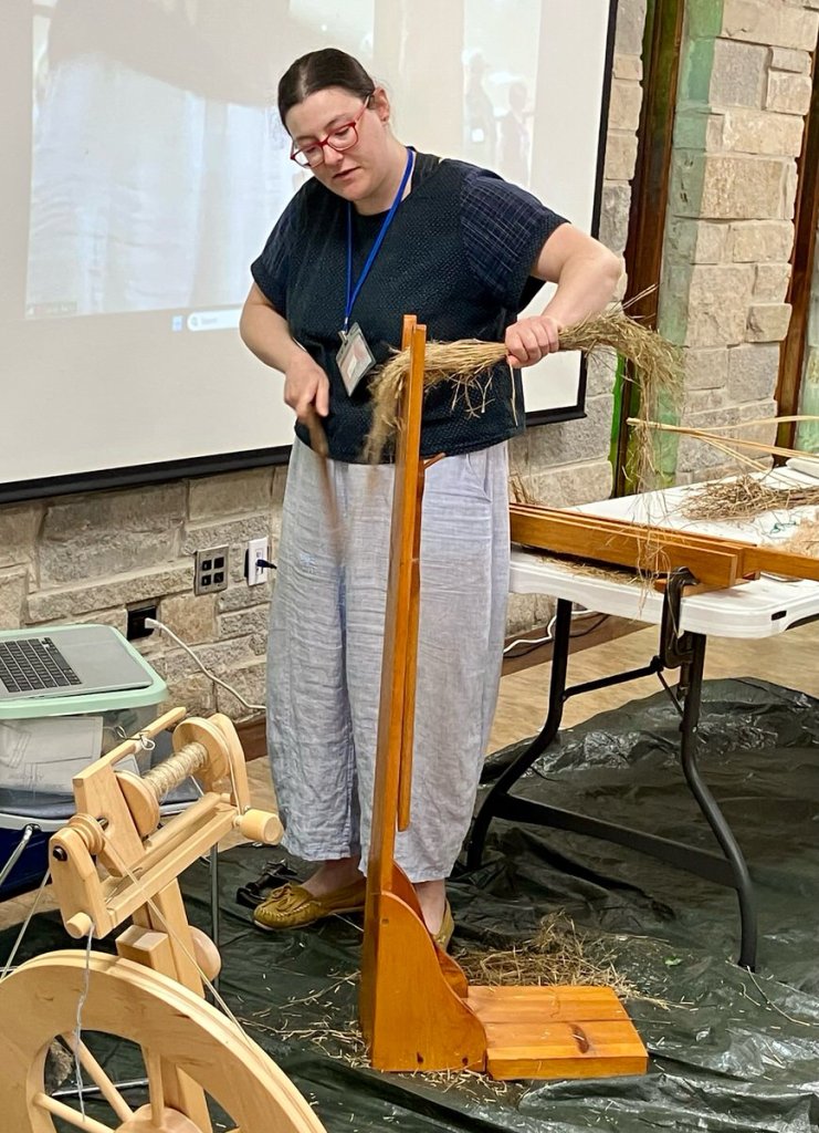 Woman standing by a tall wooden stand using a wooden tool to scrape off loose pieces of flax.