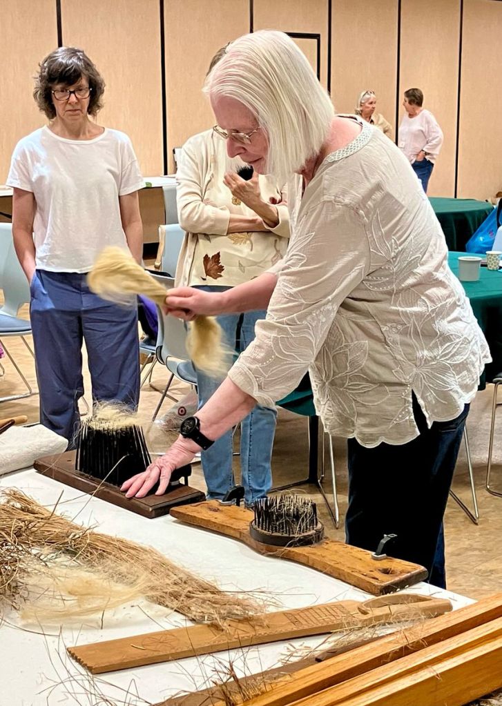 Woman standing at a table, drawing flax through a spikey implement to reveal spinnable flax fiber.