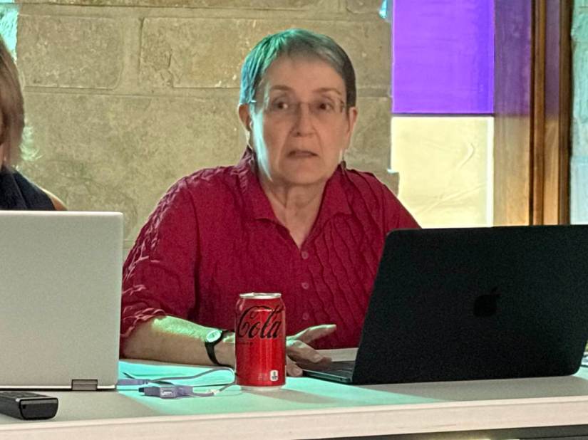 White woman with short dark hair and glasses, wearing a red shirt, sitting at a laptop computer