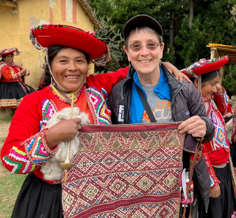 An indigenous woman wearing trditional Peruvian dress - red hat, red jacket, and black skirt, stands with a white woman wearing a brimmed hat, glasses, and a black jacket. The two women each have an arm around the other and are holding an intricately woven cloth between them with their other hands. The indigenous woman is also holding a drop spindle and wool for spinning in the hand she's holding the cloth with.