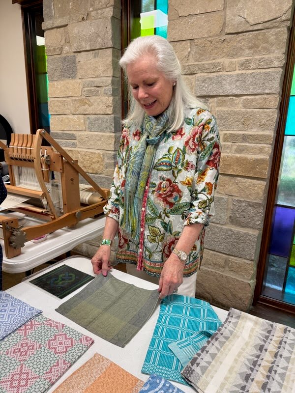A woman with long gray hair and a patterned top holds a small textile on a table.